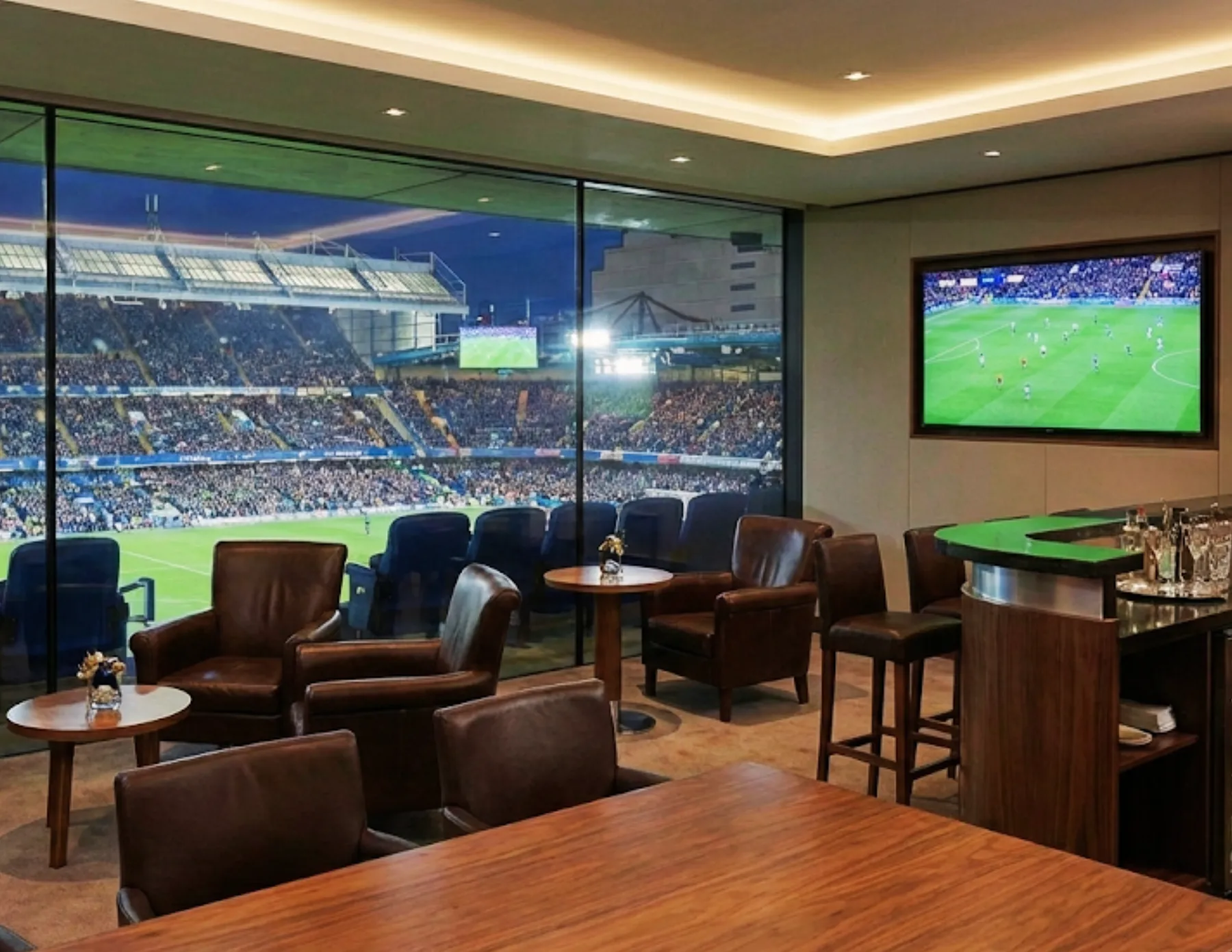 Luxury private stadium suite with lounge seating, bar area, and floor to ceiling glass overlooking a live sports event