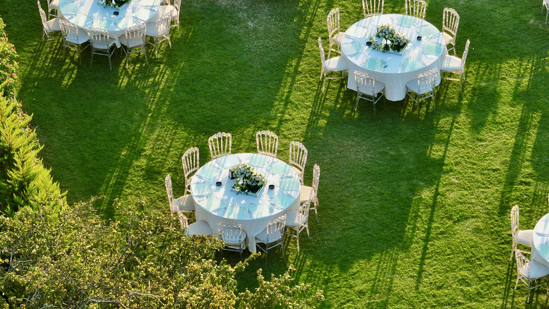 Outdoor garden event setup with round tables and elegant seating Outdoor garden event with round tables, white linens, and clear chairs arranged on a green lawn for an upscale private gathering