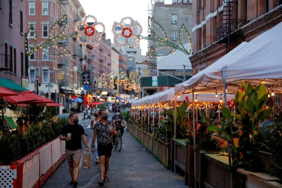 Busy city street with outdoor restaurant seating under white tents, people walking and cycling, and decorative lights hanging above the roadway at dusk.