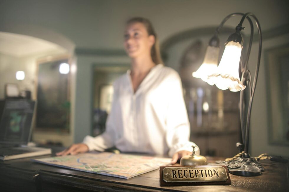 Smiling hotel receptionist standing at a classic boutique hotel front desk ready to welcome and assist arriving guests