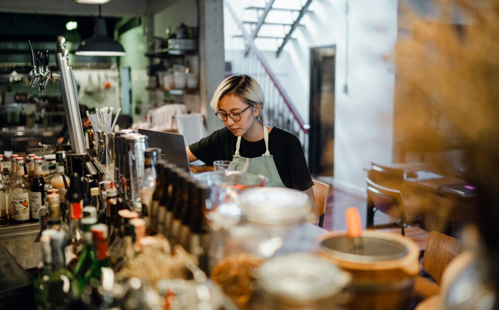 Focused restaurant staff member in apron working on a laptop at a fully stocked bar, managing operations at a professional hospitality venue