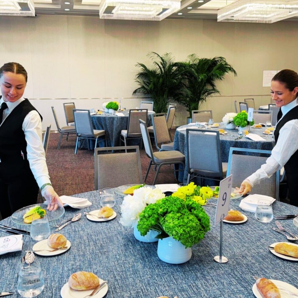 Two uniformed HMG+ catering staff professionally setting up tables at a corporate banquet hall ahead of a formal event