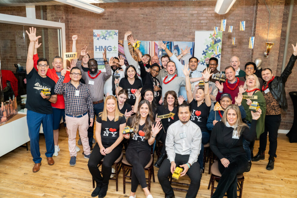 The HMG+ hospitality staffing team celebrating at their annual New York City awards event wearing I Love NY shirts in front of the HMG+ branded banner
