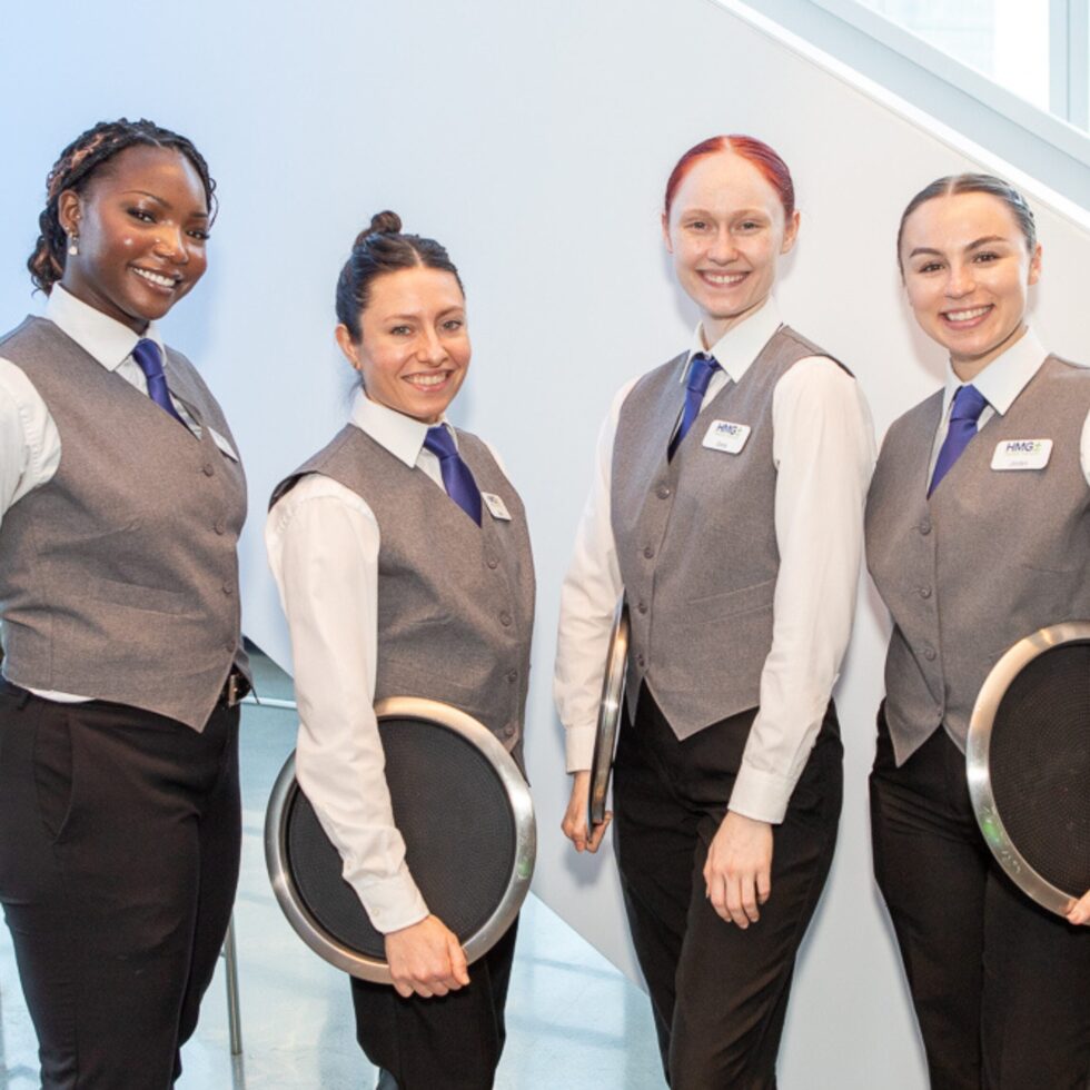 Four uniformed HMG+ professional catering staff smiling and ready to serve at a corporate event in New York or Chicago
