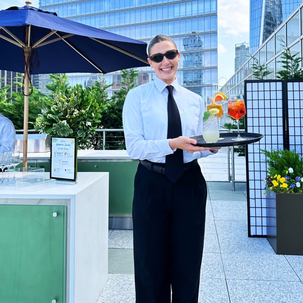 Smiling HMG+ event staff member serving cocktails on a tray at an upscale rooftop event with New York City skyline in the background