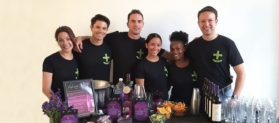 A smiling team of six event staff in matching black t-shirts with a green plus sign logo, posing together behind a bar setup featuring bottles, glassware, menus, and fresh citrus garnishes at what appears to be a promotional or hospitality event.