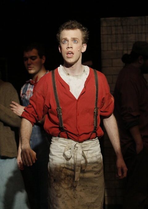 A young male actor on stage in a theatrical production, wearing a red shirt with suspenders and a dirty white apron, with an expressive, wide-eyed look on his face. Other cast members are visible in the background against a dark stage set.