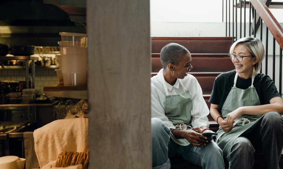 Two smiling restaurant staff members in aprons sharing a moment together near a professional kitchen at a hospitality venue