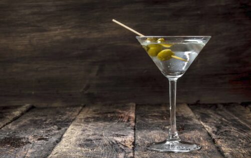 A classic martini in a chilled glass, garnished with two green olives on a cocktail pick, set on a weathered dark wooden surface.