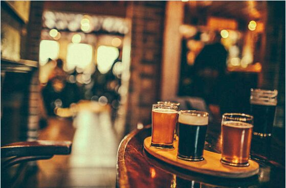 A flight of four craft beers in small glasses ranging from amber to dark stout, arranged on a round wooden serving board on a bar table, with the warm, blurred glow of a busy pub in the background.