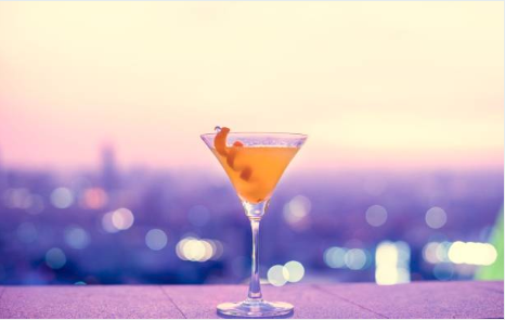 A golden cocktail in a martini glass garnished with a citrus twist, set against a soft, bokeh-lit city skyline at dusk in hues of pink and purple.