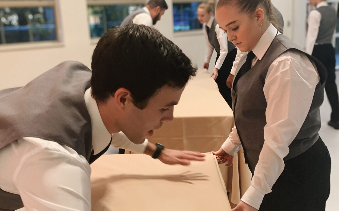Two hospitality staff members in white shirts and grey vests carefully smooth and align a beige tablecloth on a banquet table, with additional staff setting up tables in the background of a bright event space.