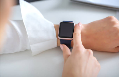 A person in a white shirt tapping the screen of a smartwatch with a navy blue band on their wrist, against a clean white desk surface.