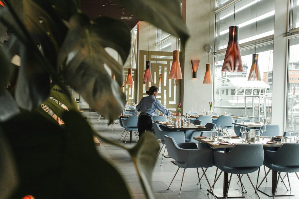 Professional restaurant staff setting elegant tables in a modern upscale dining room with designer pendant lights and waterfront views