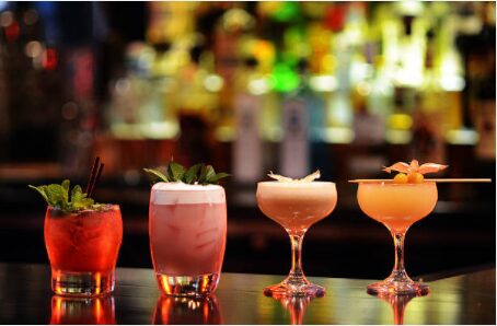 Four colorful cocktails lined up on a bar counter — a red drink with mint, a pink frothy highball, a pale pink coupe, and a coral cocktail with a fruit garnish — with a softly lit display of bottles glowing in the background.