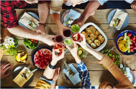 An overhead view of a group of people clinking glasses of red wine and cocktails in a toast over a rustic wooden table filled with colorful dishes, salads, roasted vegetables, fresh flowers, and shared plates.