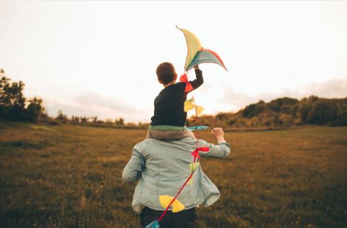 A child sitting on his father's shoulders holding a colorful kite aloft as they walk through an open grassy field, both viewed from behind against a soft, golden evening sky.