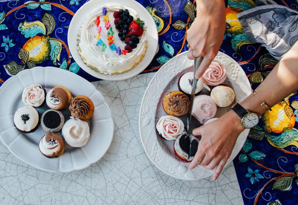 An overhead view of a dessert spread on a mosaic table with a colorful floral tablecloth. Two white plates are filled with assorted cupcakes featuring vanilla, chocolate, and pink rose-swirl frosting.