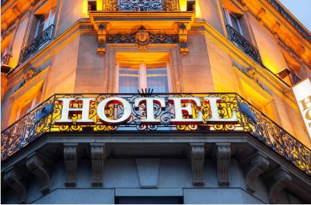 A glowing 'HOTEL' sign mounted on an ornate wrought-iron balcony of a grand, warmly lit stone building at dusk, evoking a classic European city streetscape.