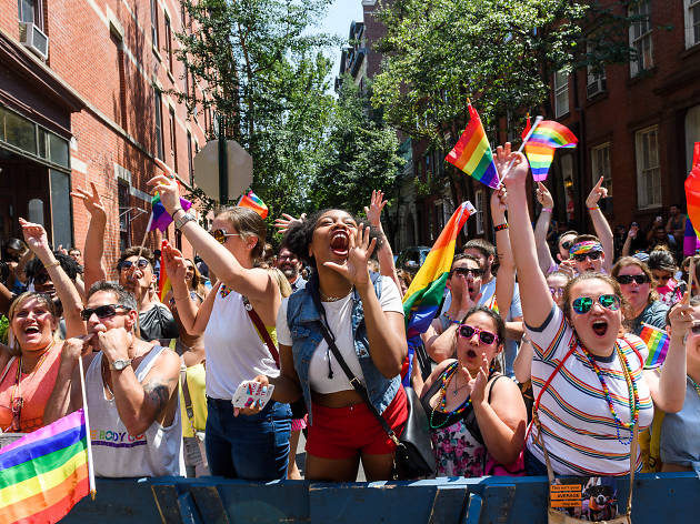 Crowd cheering and waving rainbow flags during a Pride parade on a city street.