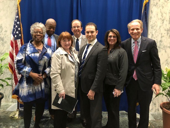 A group of seven professionally dressed adults posing together for a photo in what appears to be a government or official building, with an American flag and blue draping visible in the background.