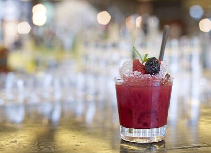 A deep red berry cocktail in a rocks glass filled with crushed ice, garnished with a blackberry, strawberry, and a metal straw, set on a gold bar counter with a softly blurred bar interior in the background.
