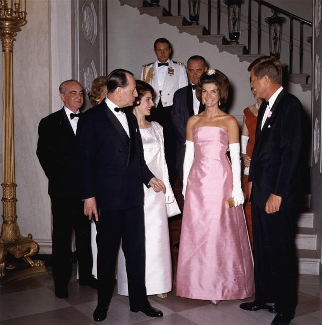A vintage color photograph of a formal gathering, showing elegantly dressed guests in tuxedos and evening gowns socializing near an ornate staircase in what appears to be a grand official venue, with a uniformed military officer visible in the background.