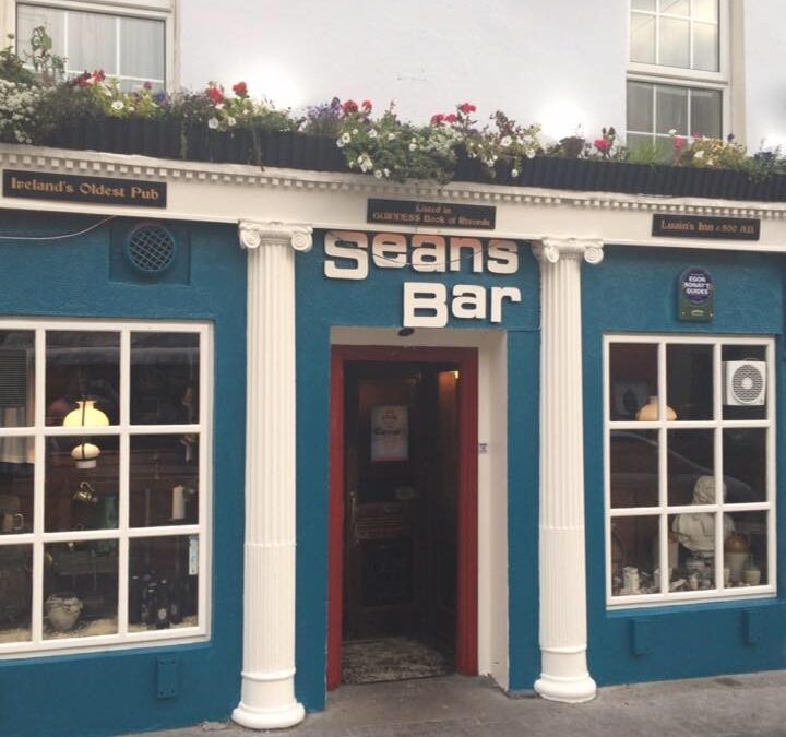 The exterior of Sean's Bar, a teal-painted pub with white columns, a red door, and flower boxes along the roofline. Signage identifies it as 'Ireland's Oldest Pub,' listed in the Guinness Book of Records.