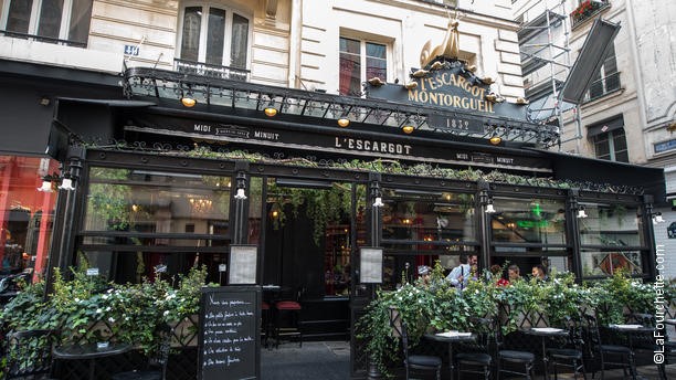 The classic black façade of L'Escargot Montorgueil, a historic Parisian restaurant established in 1832, featuring an ornate storefront with lush greenery, a sidewalk terrace with outdoor seating, and a chalkboard menu on the street.