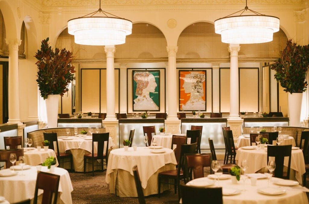 An upscale restaurant dining room with white linen-draped tables, dark wood chairs, large drum pendant lights, and arched columns, accented by bold colorful portrait paintings on the wall and tall floral arrangements in white vases.