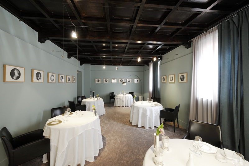 An intimate fine dining room with round tables draped in white linen, black leather chairs, a dark coffered ceiling, and pale blue-grey walls lined with framed portrait artwork, with sheer curtained windows letting in soft natural light.