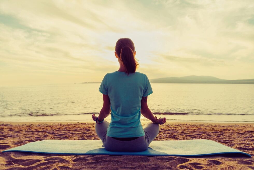 A woman with a ponytail sits in a cross-legged meditation pose on a yoga mat facing the ocean, viewed from behind, against a warm golden sunset sky with distant hills on the horizon.