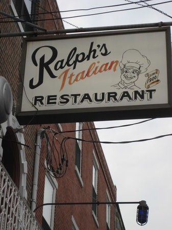 An outdoor sign for Ralph's Italian Restaurant, featuring a illustrated chef logo and 'Since 1900' badge, mounted on a brick building under an overcast sky.