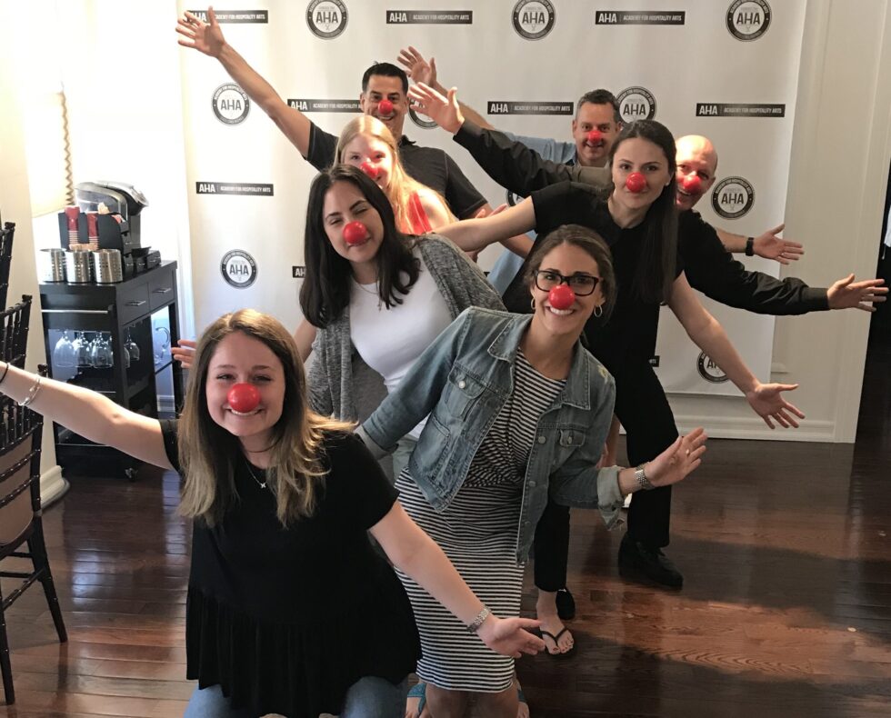 A cheerful group of seven people wearing red clown noses strike fun, playful poses in front of an AHA (Academy for Hospitality Arts) branded step-and-repeat backdrop, with a bar service cart visible to the left.