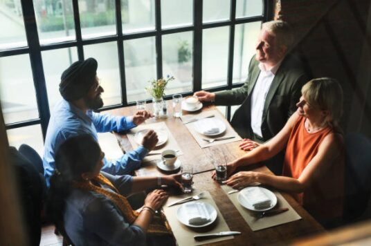 An overhead view of four adults engaged in conversation around a rustic wooden dining table set with plates, wine glasses, and coffee cups, beside large windows in a warmly lit restaurant.