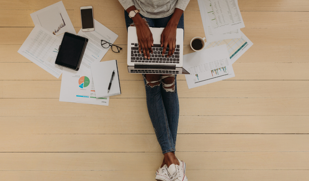 An overhead view of a person in ripped jeans and sneakers sitting on a light wood floor, typing on a laptop surrounded by printed charts, spreadsheets, a tablet, smartphone, glasses, a pen, and a cup of coffee.