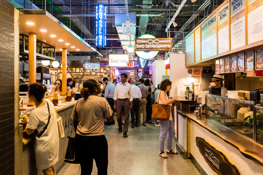 A bustling food market scene with food stalls lined on either side of the aisle, people buying food at the stalls and people walking up and down the aisle