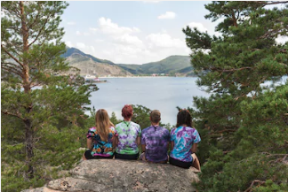 Four people wearing colorful tie-dye shirts sit side by side on a rocky ledge, viewed from behind, overlooking a calm mountain lake surrounded by pine trees and hills under a partly cloudy sky.