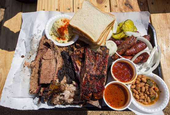 A generous Texas-style BBQ spread on a butcher paper-lined tray, featuring sliced brisket, pork ribs, sausage links, white bread, pickles, onions, coleslaw, pinto beans, and two cups of barbecue sauce, served on a wooden picnic table.