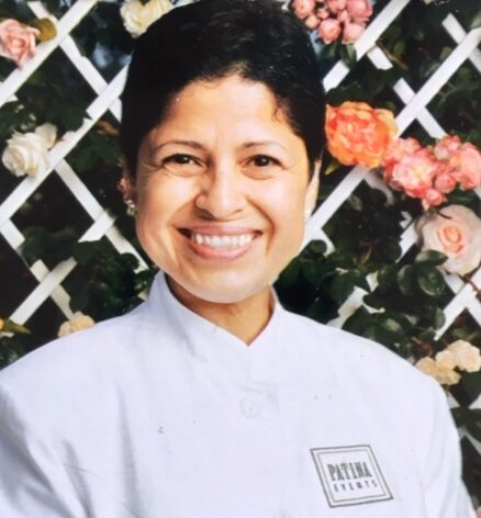 A smiling woman in a white Patina Events chef's coat poses in front of a white lattice trellis adorned with pink and peach roses.