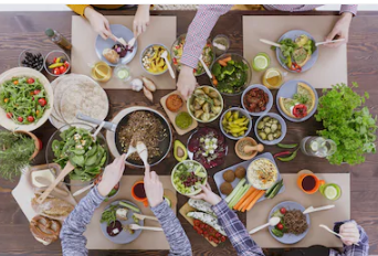 An overhead view of a vibrant shared meal, with multiple hands reaching across a wooden table filled with a colorful spread of salads, grain bowls, vegetables, dips, and fresh greens