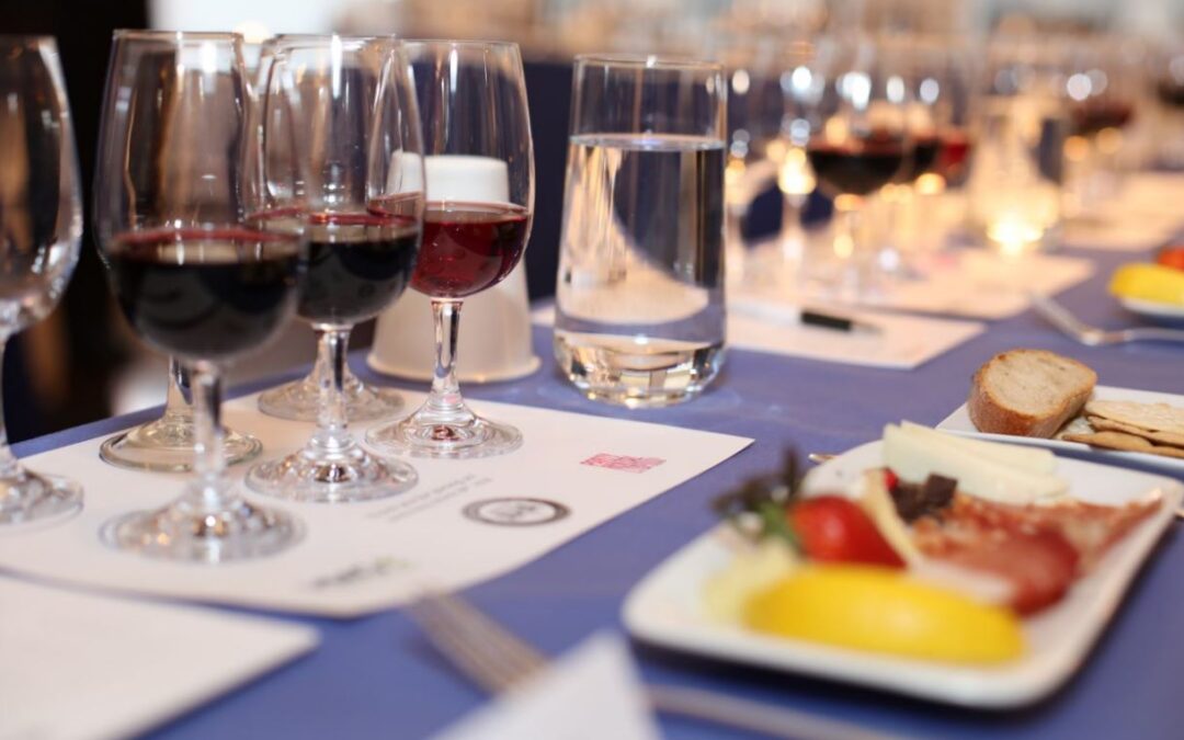 An elegantly set wine tasting table with multiple glasses of red wine, a water glass, and a printed tasting guide on a blue tablecloth. A white plate in the foreground holds an assortment of cheeses, crackers, tomatoes, and charcuterie, with more place settings visible down the length of the table.